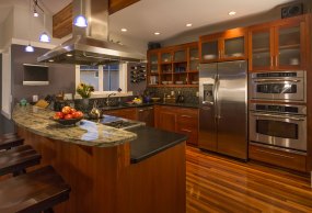 Contemporary upscale home kitchen interior with wood cabinets and floors, granite countertop, accent lighting, television &amp; stainless steel appliances including double oven, refrigerator and vent hood